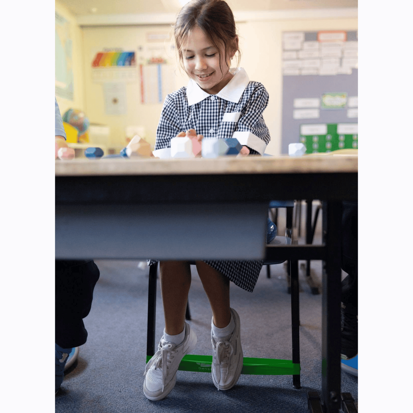 Young girl sitting at a desk in a classroom, smiling, with fidget band on chair