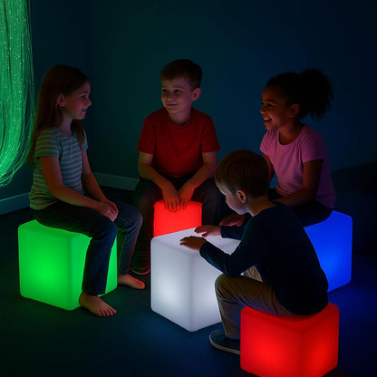 Four children sitting on glowing 40 LED cube-shaped floor lamps in a dark room, with the cubes emitting green, red, white, and blue light.