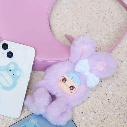 A fluffy pastel purple bunny-shaped phone case with a white bow and a small anime-style face, shown styled on a beige stone surface next to a pink bag and a white phone.
